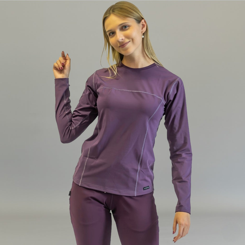 Woman wearing purple long sleeve underscrub, standing with right arm raised slightly.