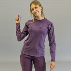 Woman wearing purple long sleeve underscrub, standing with right arm raised slightly.